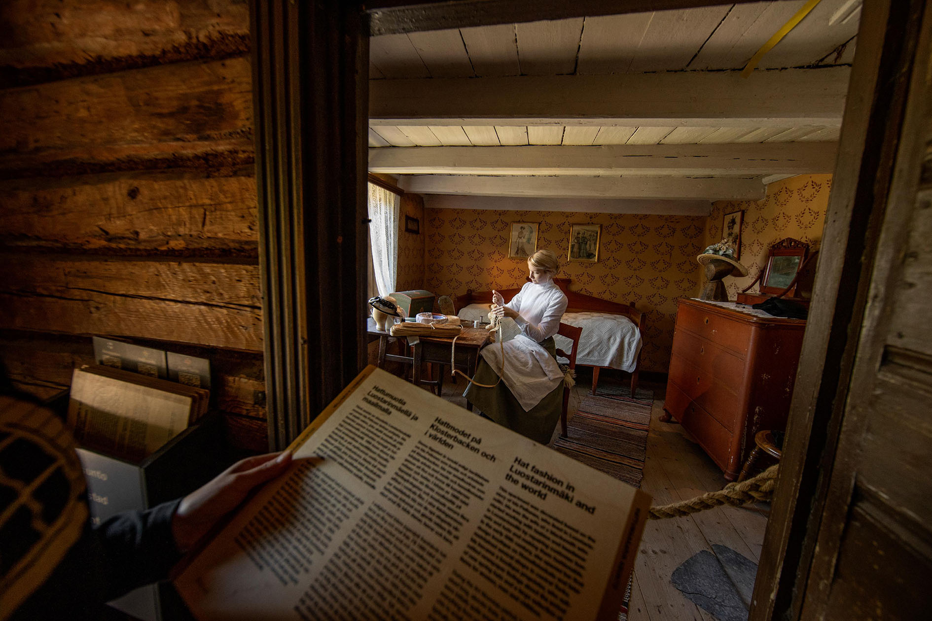 1900-luvun alkupuolen asuun pukeutunut henkilö istuu pöydän äärellä tekemässä hattua. Huone on sisustettu vanhoilla huonekaluilla. Ovensuussa museovieras lukee tilaa esittelevää tekstiä.