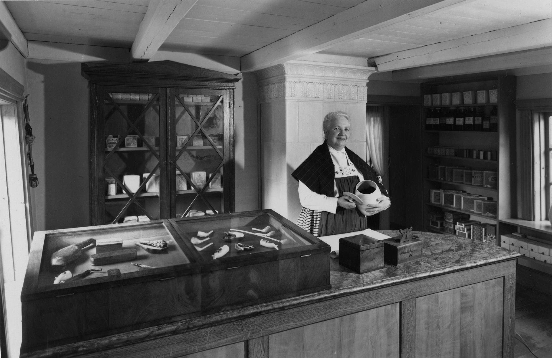 Ett gammalt foto av en museivakt i tidstypisk dräkt, som presenterar föremål i tobaksgården.